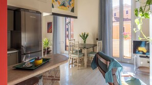 Une cuisine moderne dotée d’un réfrigérateur en acier inoxydable, un coin repas avec une table et des chaises, et un balcon offrant une vue sur les immeubles.