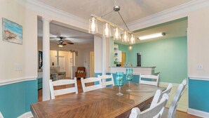 A dining area with a wooden table and white chairs, a ceiling fan, and a kitchen in the background.