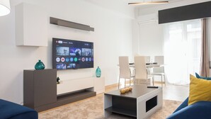 Un soggiorno moderno con una TV a schermo piatto fissata al muro, un tavolino da caffè con oggetti decorativi, un divano e una zona pranzo con sedie.