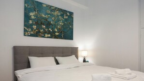 Un letto rifatto con lenzuola bianche e due cuscini, una lampada da comodino e un quadro appeso al muro raffigurante un albero.