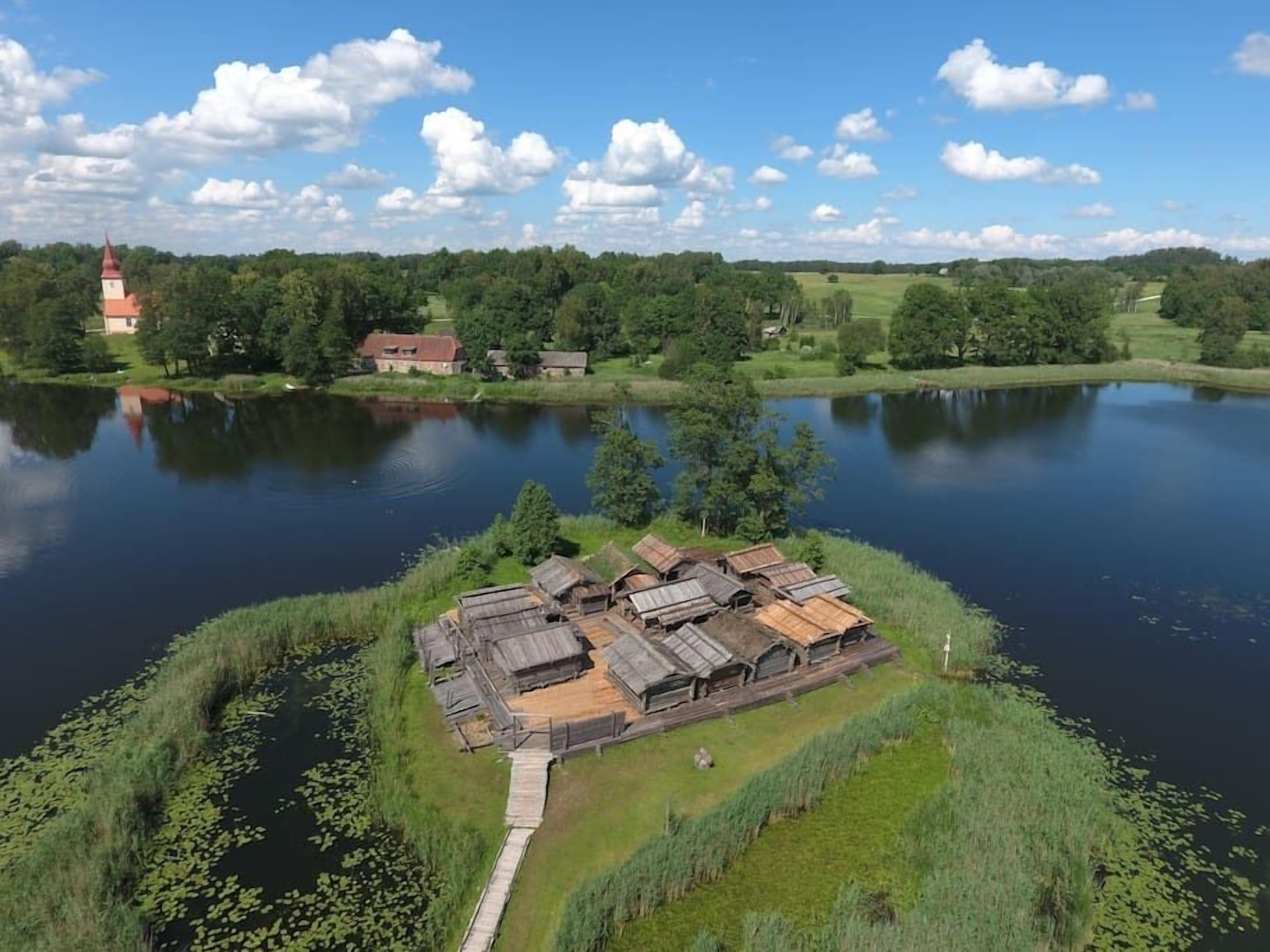 Une construction en bois au bord du lac, accessible par un sentier, entourée d’une végétation luxuriante et avec, au loin, un bâtiment dont le toit est rouge.