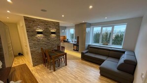 A modern living room with a dining area, a stone accent wall, and a large window with a view of trees. - Bright and modern apartment in Cesis, Luxurious  stay at modern apartment (Cesis) (Cēsis)