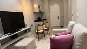 Una sala de estar moderna con televisor de pantalla plana, un sofá con cojines, una mesa de comedor con sillas y una cocina con armarios y una lámpara colgante.
