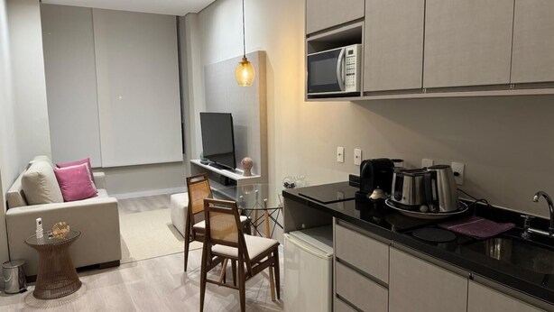 Una cocina moderna con microondas integrado, cafetera y hervidor en la encimera. Hay una zona de comedor con mesa de vidrio y sillas, y un sofá con cojines.