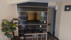 Een woonkamer met een flatscreen televisie aan een donkere muur, een plant en een tv-meubel met planken.