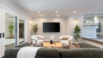 A modern living room with a sofa set, a coffee table, and two armchairs. A flat-screen TV is mounted on the wall, and there are potted plants on either side.