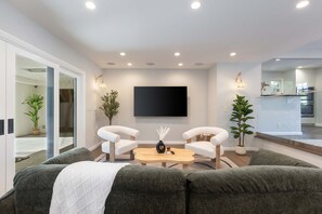 Un salon moderne avec un canapé, une table basse et deux fauteuils. Un téléviseur à écran plat est fixé au mur, et de chaque côté, on trouve des plantes en pot.