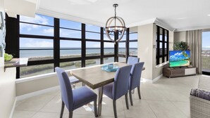 A modern living room with a large window overlooking a scenic view, a dining area with a table and chairs, and a television on a wooden stand. - Front Beachfront Condo | Pool, Fitness & Grills (Marco Island)