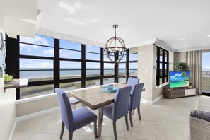 A modern living room with a large window overlooking a scenic view, a dining area with a table and chairs, and a television on a wooden stand. - Front Beachfront Condo | Pool, Fitness & Grills (Marco Island)