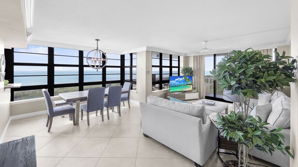 A modern living room with a large window, a dining area, and a sofa. - Front Beachfront Condo | Pool, Fitness & Grills (Marco Island)