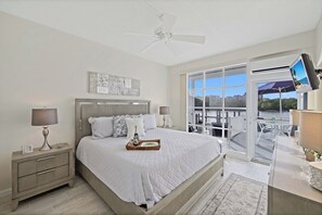 A bedroom with a large bed, a nightstand, a ceiling fan, a TV mounted on the wall, and a view of the outdoors through a sliding glass door. - Coastal waterfront condo located in Olde Marco (Marco Island)
