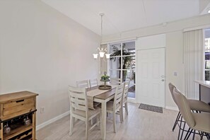 A dining area with a wooden table, white chairs, a chandelier, and a view through a door to the outside.
