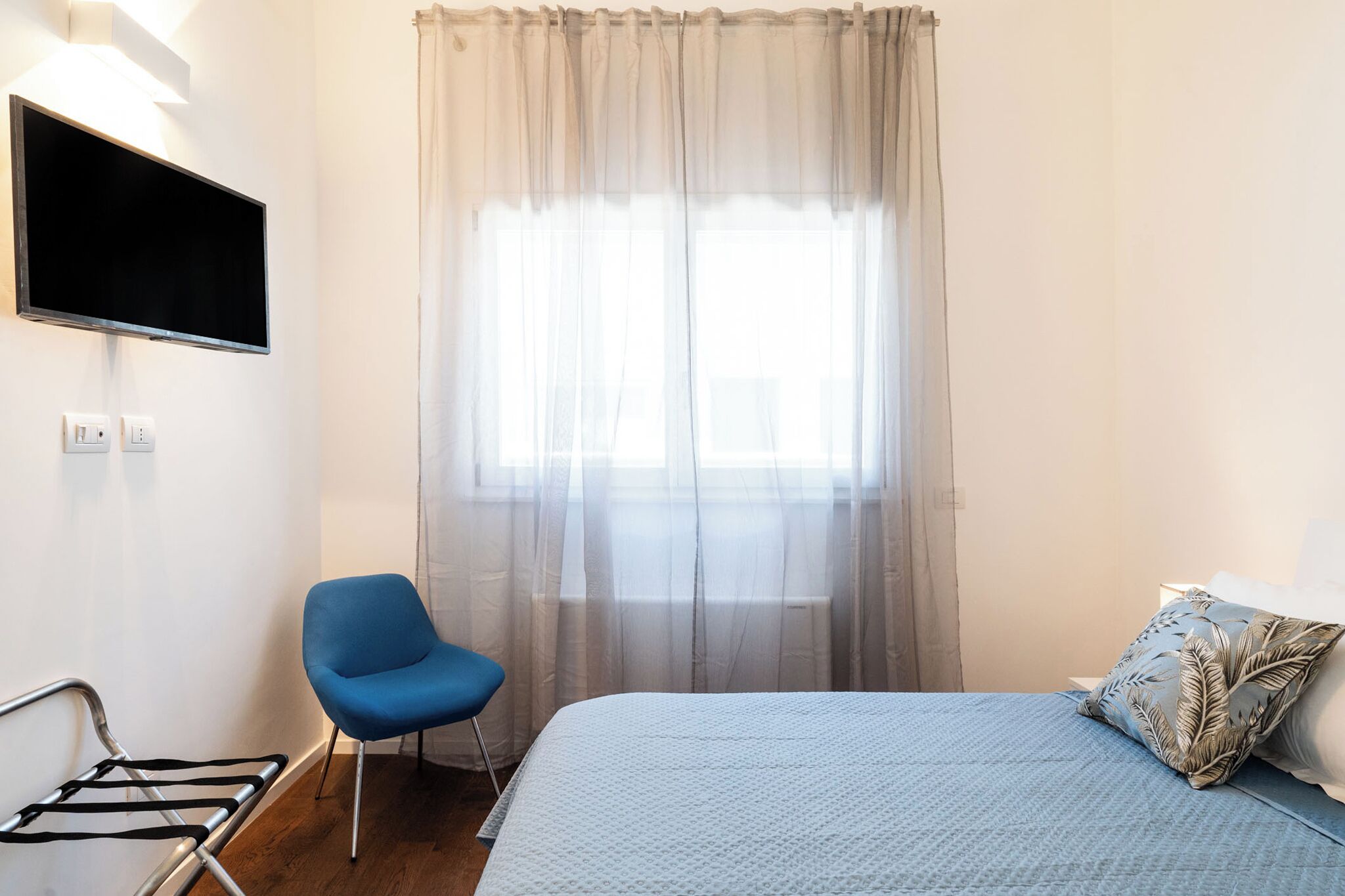 Un dormitorio con una cama, una silla azul, un televisor montado en la pared y una ventana con cortinas translúcidas.