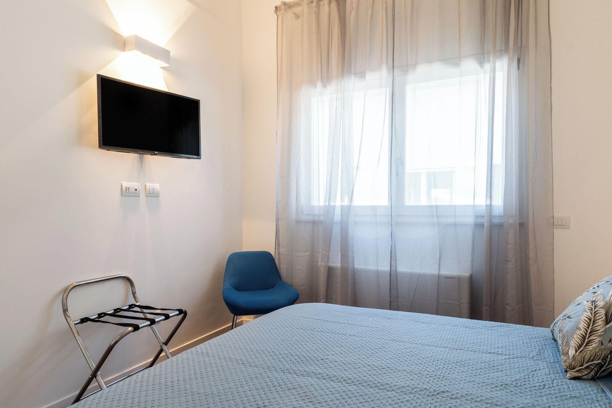 A hotel room with a bed, a blue chair, a TV mounted on the wall, and a window with sheer curtains.