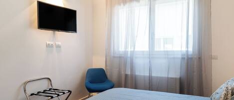 A hotel room with a bed, a blue chair, a TV mounted on the wall, and a window with sheer curtains.