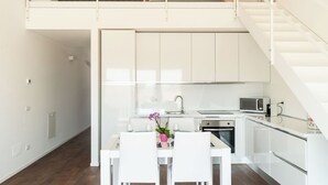 Une cuisine moderne avec des meubles blancs, une table à manger avec des chaises et un escalier menant à une chambre située en mezzanine.