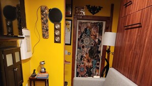 Un salón con un sofá gris, un mueble de madera, una mesita con una licuadora, una alfombra colorida y una puerta con papel tapiz floral.