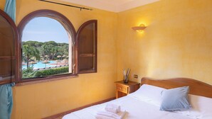 A hotel room with a bed, a nightstand, a window with a view of a pool and greenery, and a wall-mounted light.