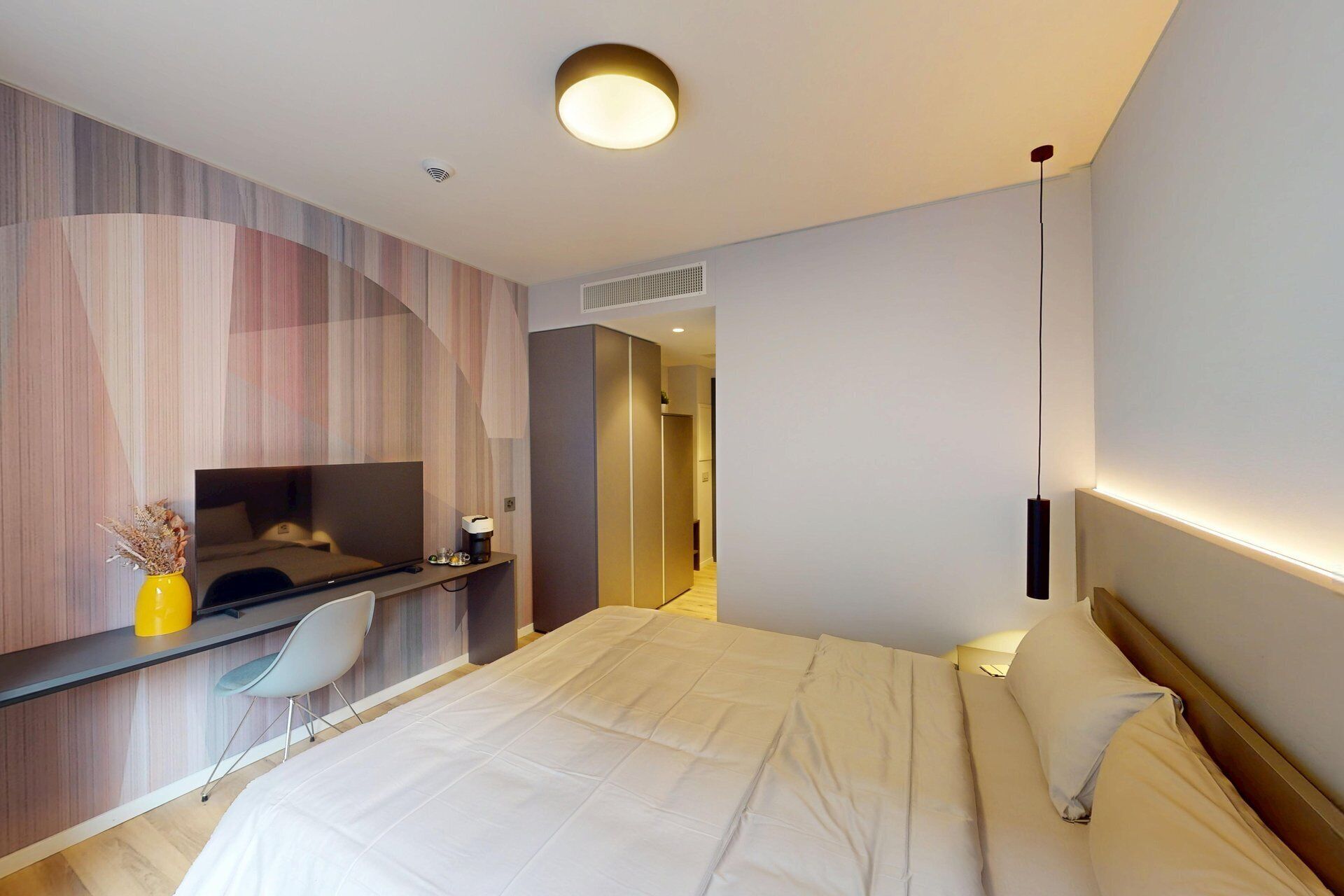 Ein modernes Hotelzimmer mit einem Bett, einem Schreibtisch mit Stuhl, einem Fernseher und einem Nachttisch mit Lampe.