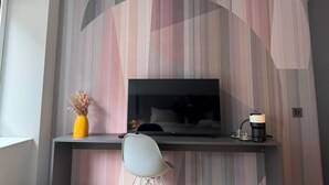 Een moderne woonkamer met een flatscreen televisie die aan een muur met een strepenpatroon is bevestigd. Er staat een kleine tafel met een vaas gedroogde bloemen, een koffiezetapparaat en een glas. Een witte stoel is voor de tafel geplaatst.