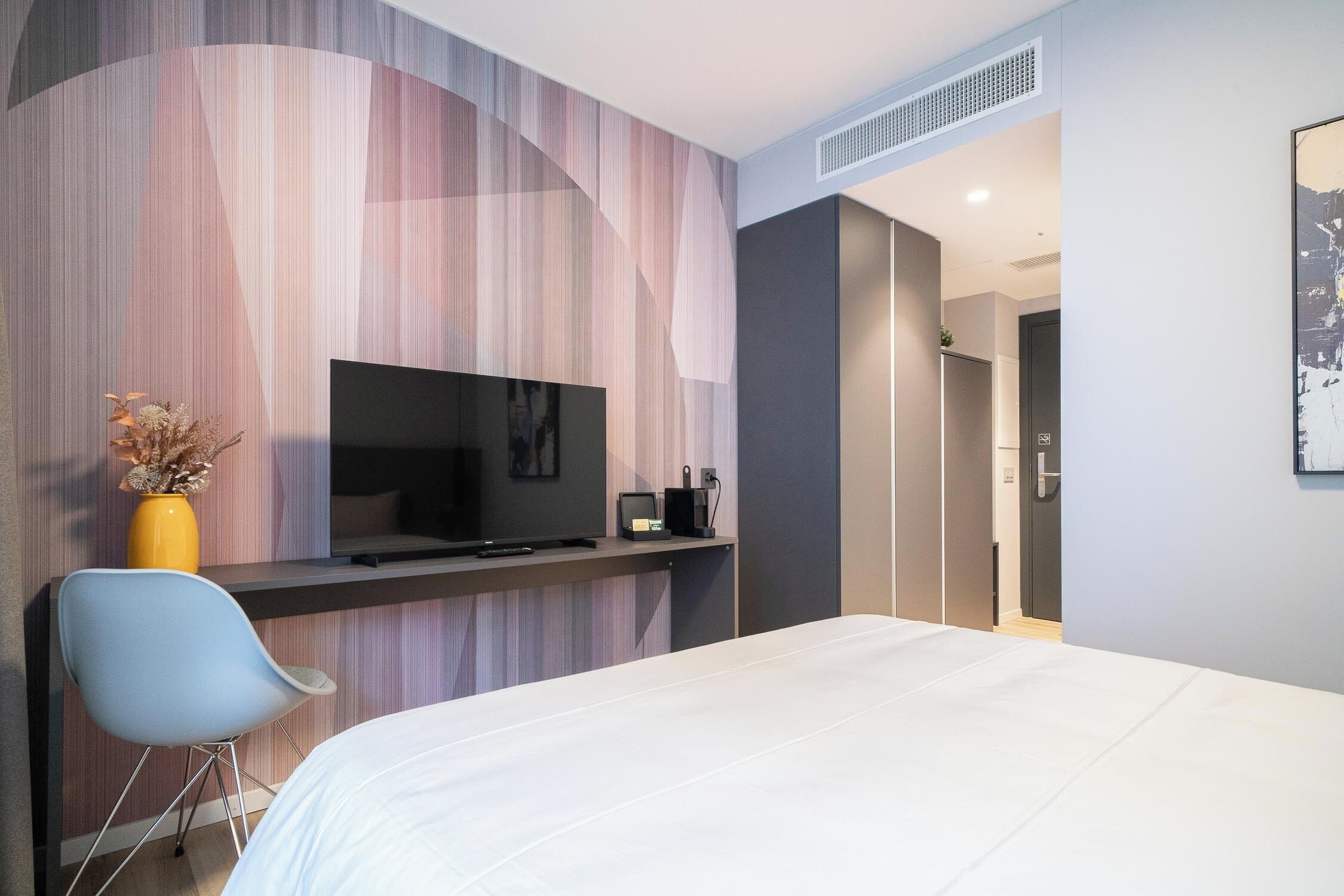 Une chambre d’hôtel moderne, équipée d’un lit, d’un téléviseur posé sur un meuble, d’un bureau avec une chaise et d’un mur orné d’un motif à rayures.