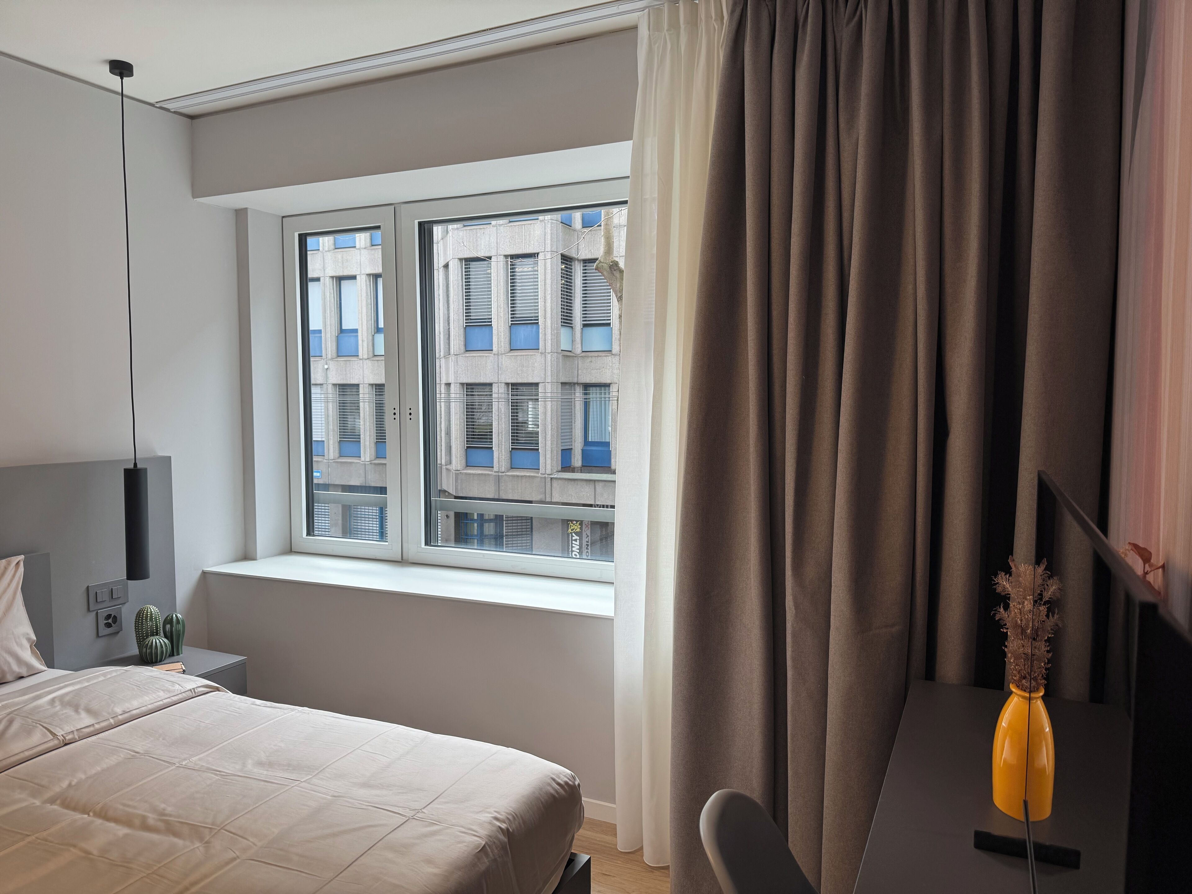 A modern hotel room with a large bed, a desk with a chair, a window with blinds, and a vase with dried plants.
