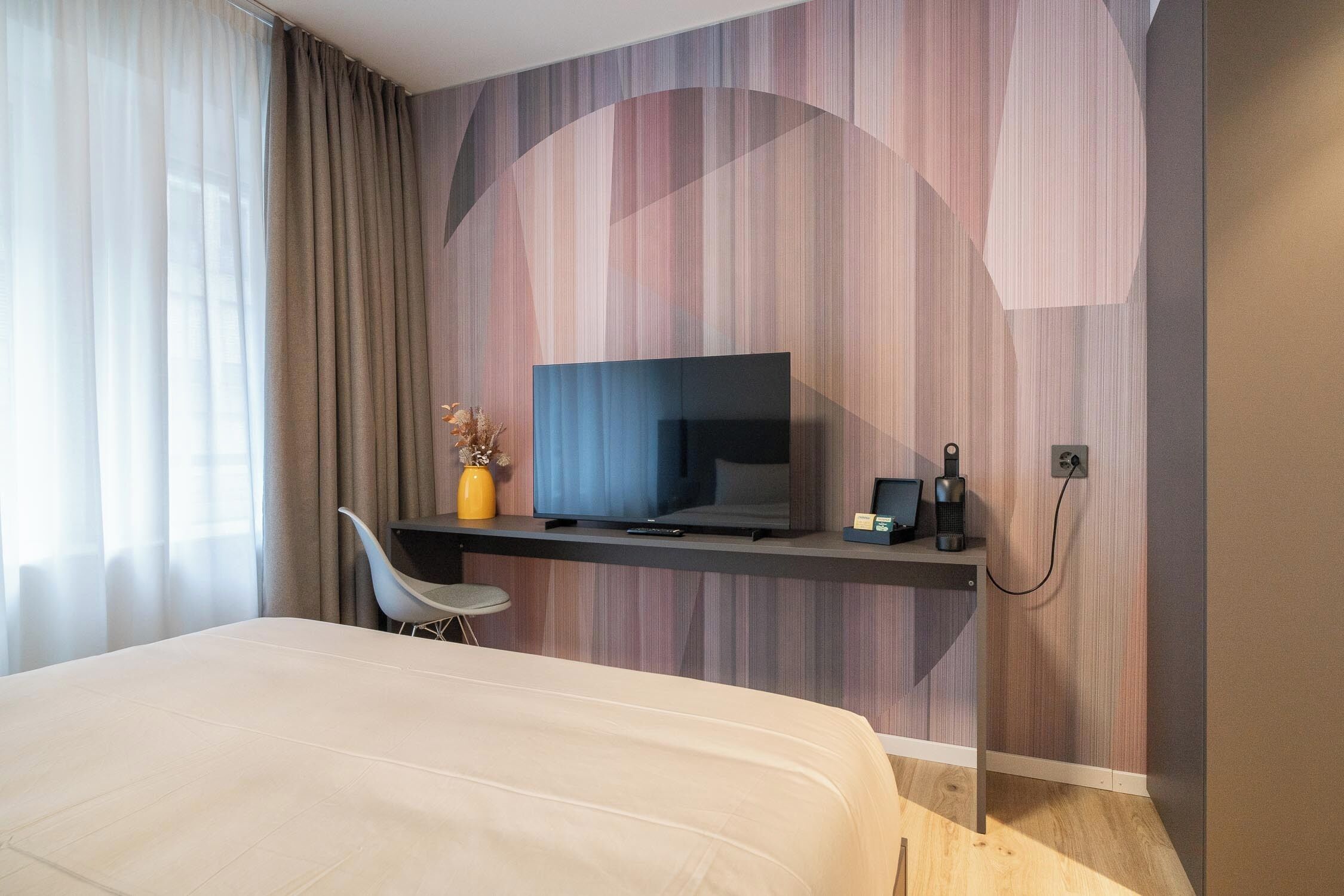A modern hotel room with a large bed, a TV mounted on a wall, a desk with a chair, and a vase with flowers.