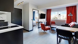 Una habitación de hotel moderna con kitchenette, un televisor de pantalla plana, un comedor con mesa y sillas, y un ventanal con cortinas.