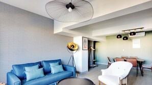 Una sala de estar moderna con un sofá azul, una mesa de centro redonda y un sillón blanco. Se observa un comedor con una mesa y sillas al fondo.