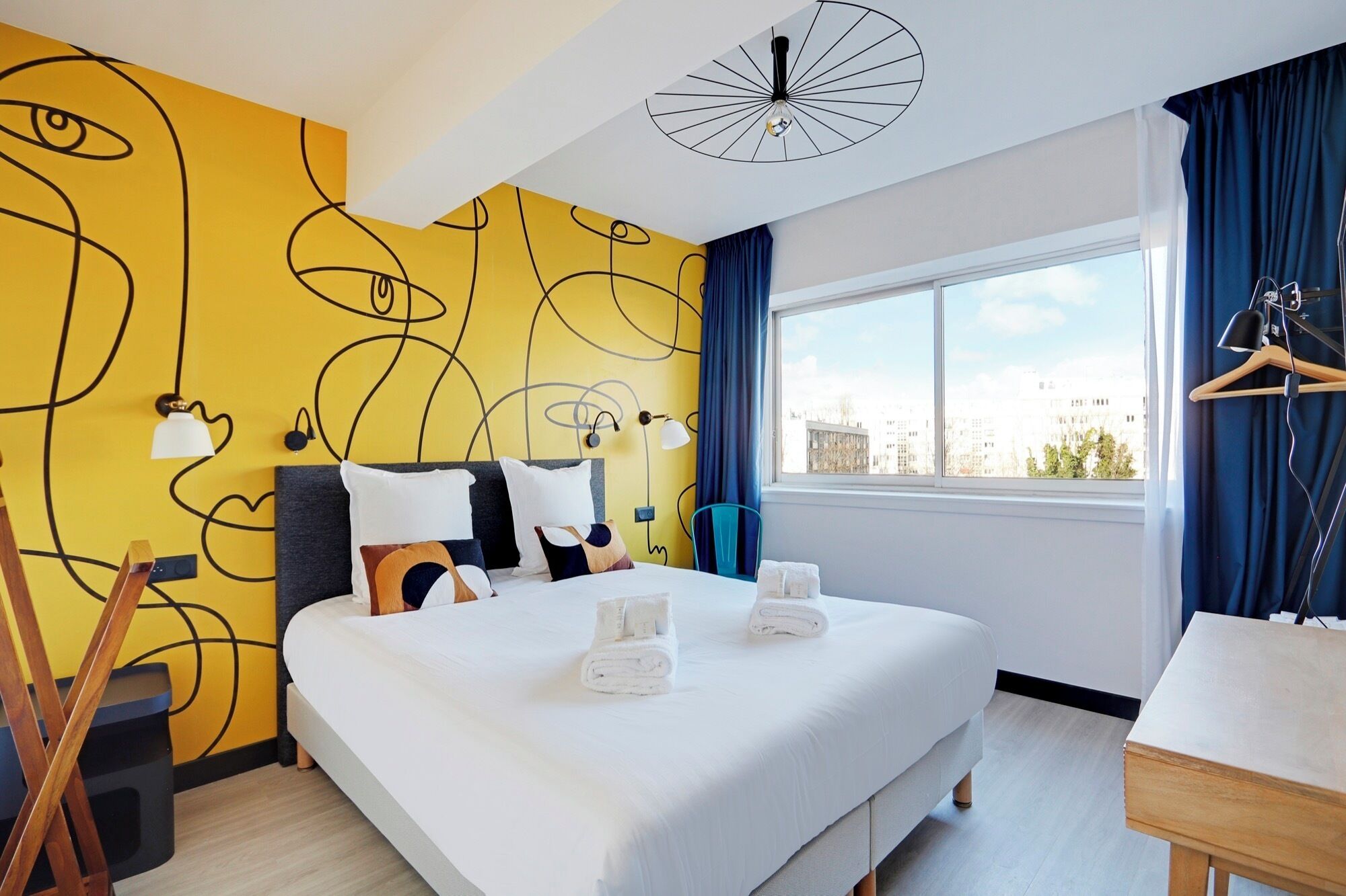 A hotel room with a large bed, a view of the city, and a unique yellow wall design.