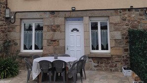 Un edificio de dos pisos con fachada de piedra y techo plano. Cuenta con una puerta blanca con un pequeño ventanal encima, y dos ventanas en el nivel superior. Frente al edificio, hay una mesa con sillas y un pequeño jardín con plantas.