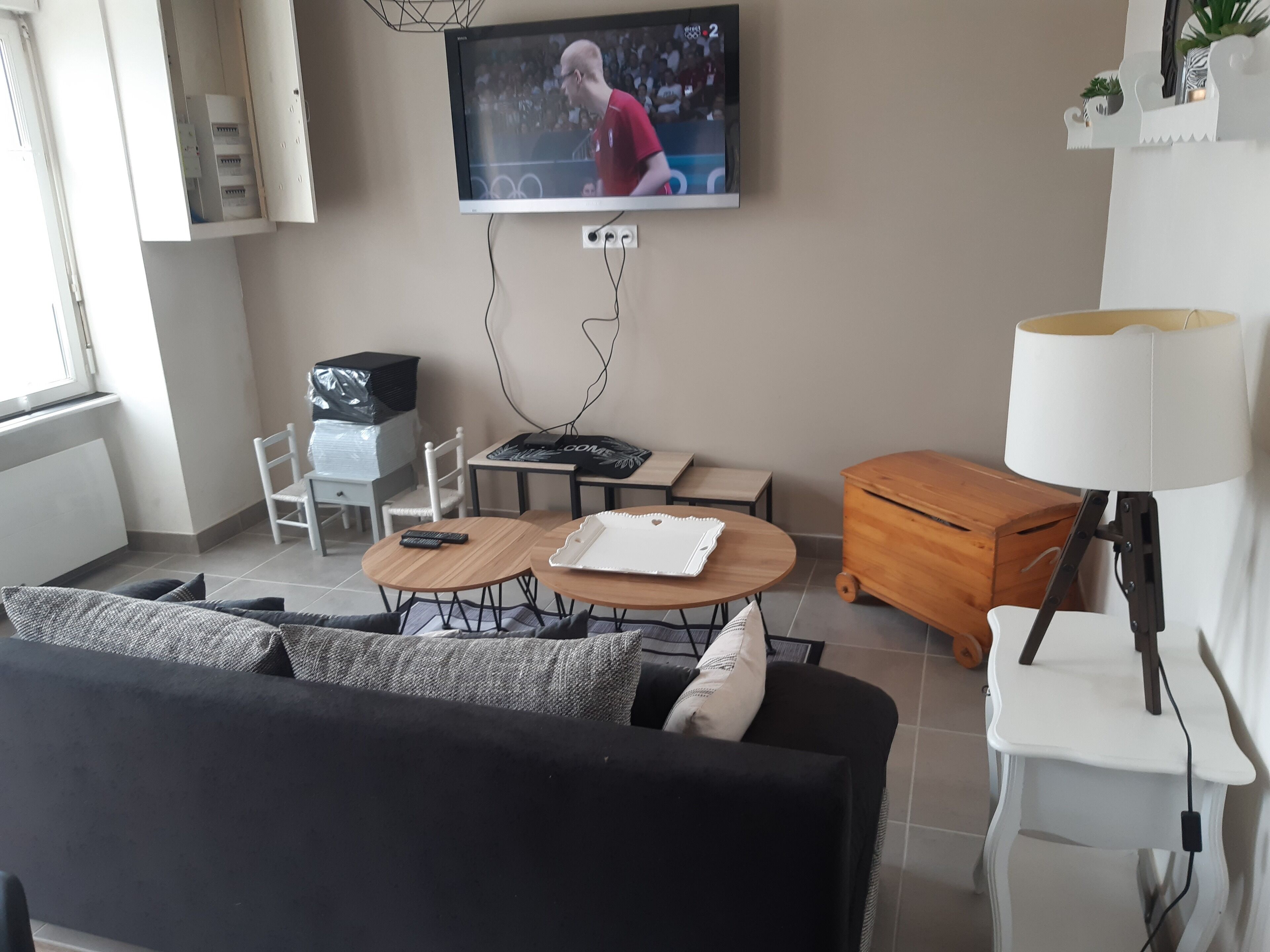 Een woonkamer met een aan de muur bevestigde televisie, een bank, een salontafel, een lamp en een klein houten kastje.