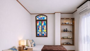 Una camera d'albergo con due letti, una finestra a vetrata colorata, uno scaffale con oggetti decorativi e un condizionatore d'aria.
