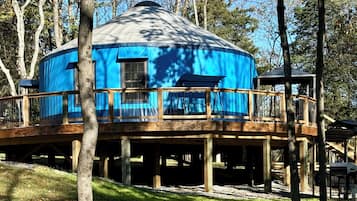 A blue yurt with a wooden deck and a small window, surrounded by trees.