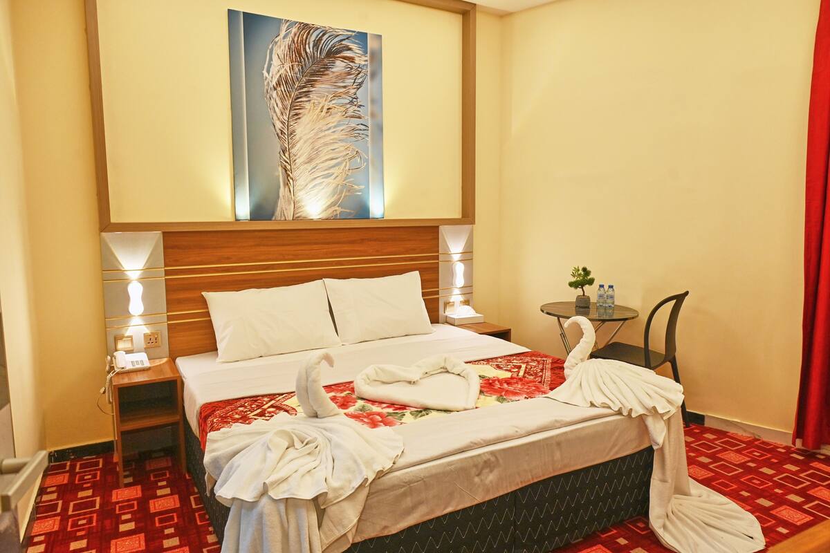 A hotel room with a neatly made bed, a small table with a plant and bottles, and a painting above the bed.