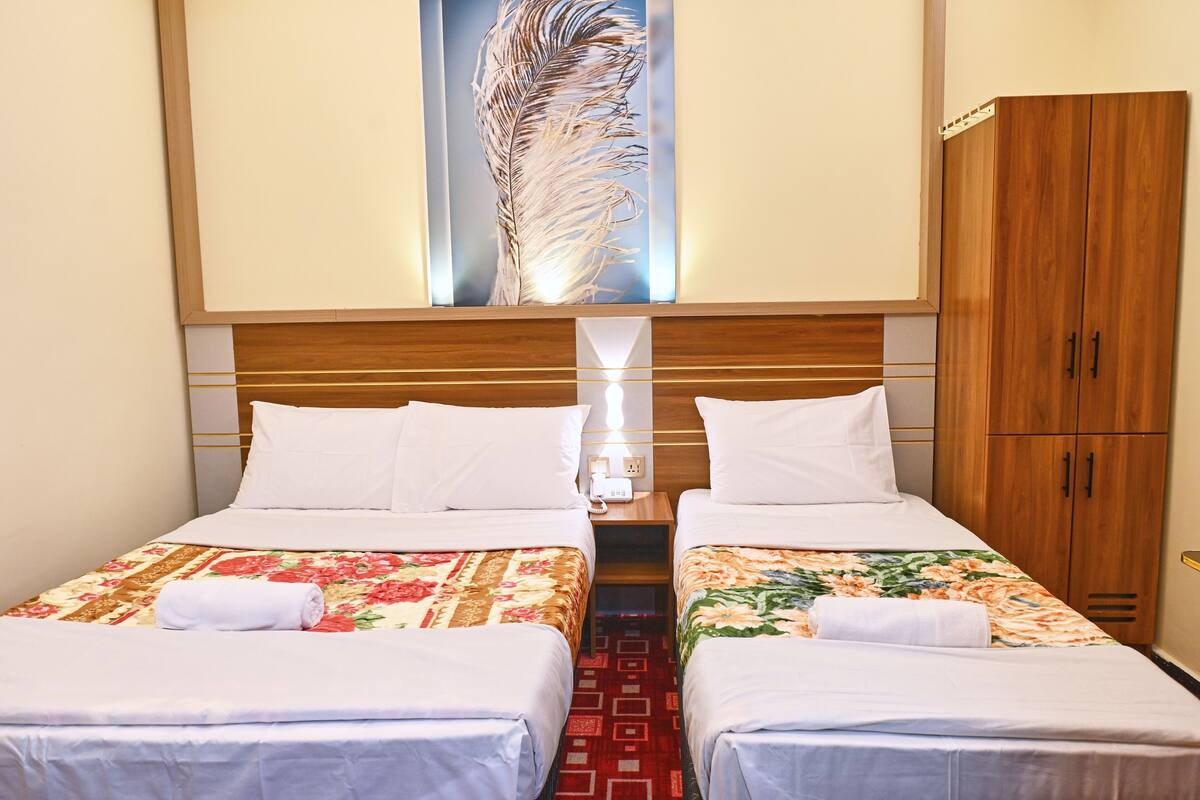 A hotel room with two single beds, a wooden wardrobe, a nightstand with a lamp, and a large feather artwork on the wall.