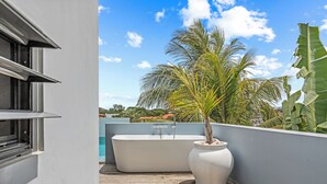 Ein moderner Balkon mit großem Pflanzgefäß, freistehender Badewanne und Blick auf Palmen und Gebäude im Hintergrund.