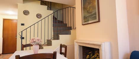A dining area with a table set for two, a vase of flowers, a fireplace with sunflowers, a staircase, and a painting on the wall.