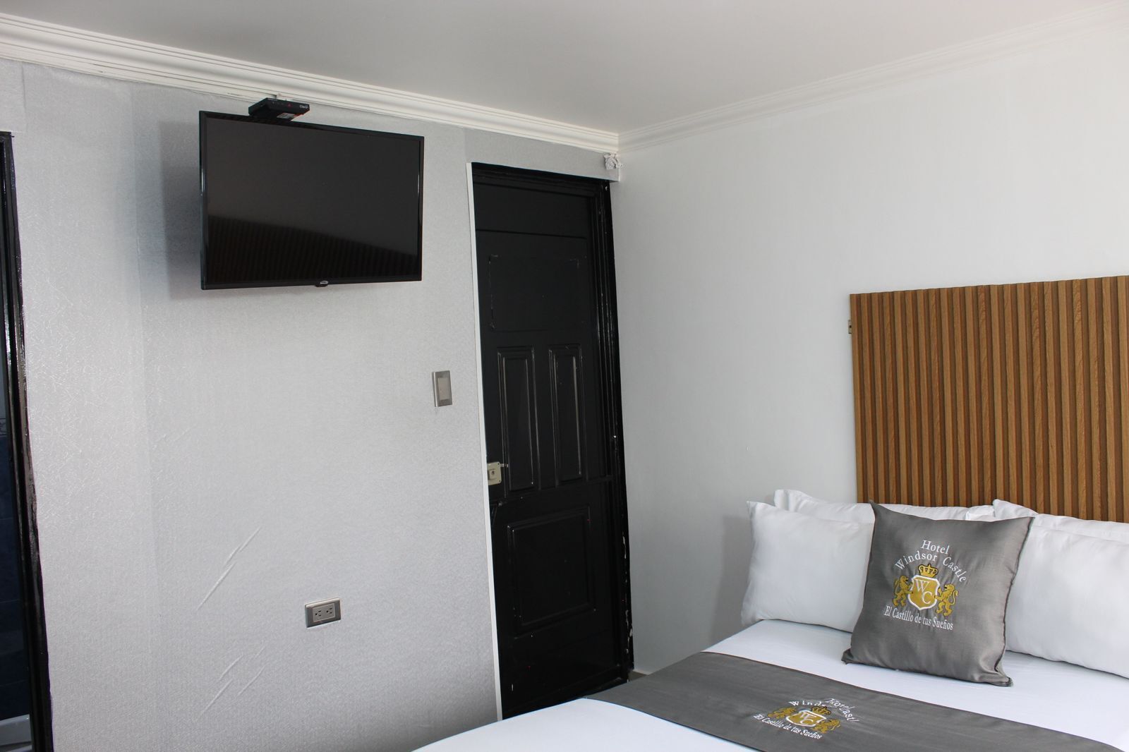 Habitación de hotel con una cama, un televisor de pantalla plana montado en la pared, una puerta negra y un cabecero de madera.