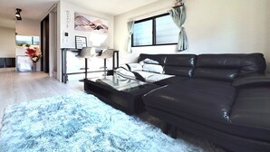 A modern living room with a black leather sofa, a glass coffee table, and a blue shag rug. There is a dining area with a bar and chairs visible in the background. - NEW! [Built in 2024, Entire Newly Built House for Rent / Free Parking Available] Experience Osaka’s Old Town! Conveniently located just 8 minutes by train from Namba! Excellent access to Osaka, Kyoto, and Nara! (大阪市)
