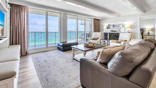 A spacious living room with a large window overlooking the ocean, a leather sofa, a coffee table, and a dining area with a chandelier.