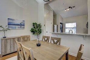 Ein Essbereich mit Holztisch und Stühlen, einer Vase mit Grünpflanzen, einem Bild an der Wand und einer Küchenzeile mit Kühlschrank und Spüle.