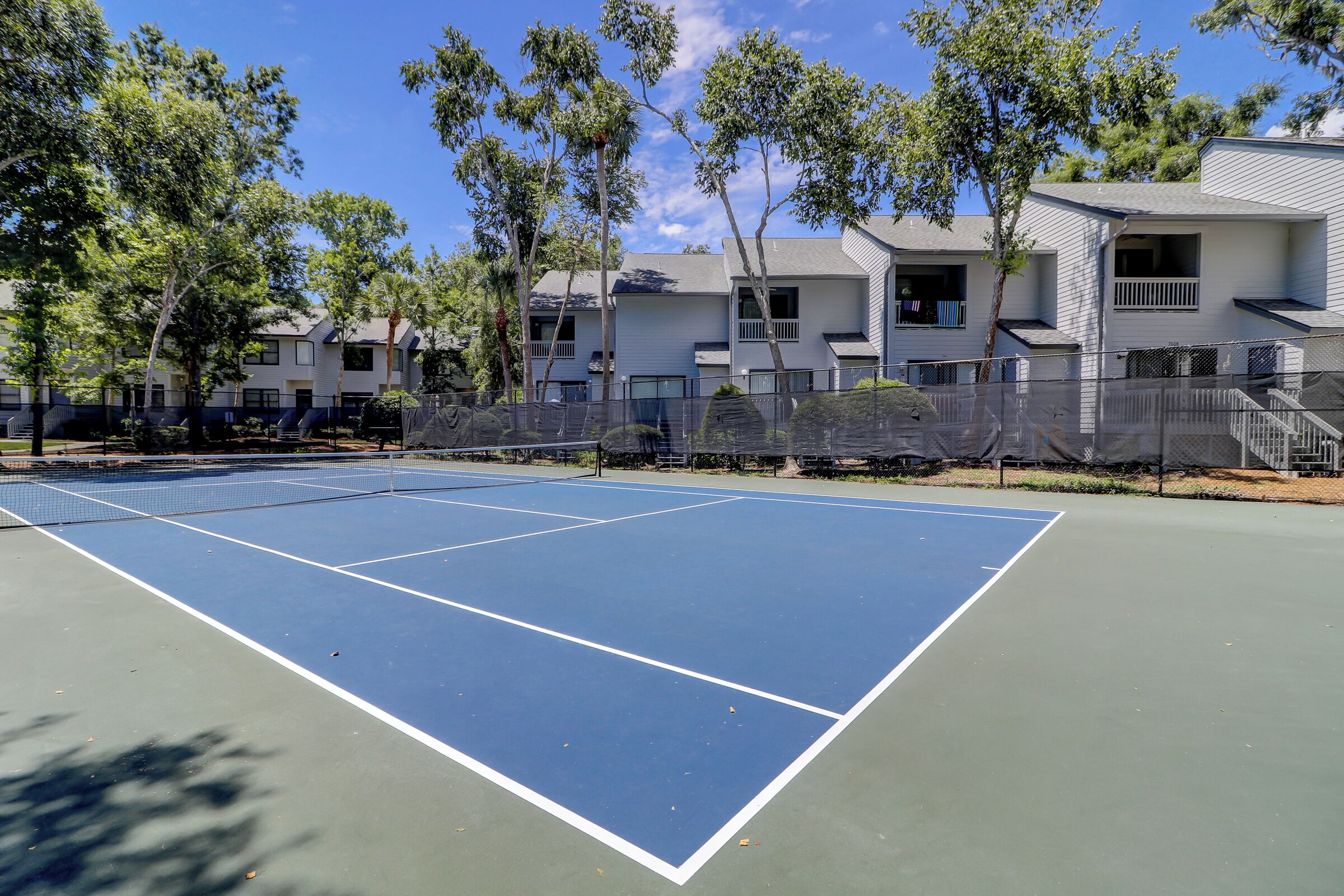 Una cancha de tenis con red, rodeada de árboles y un edificio con balcones.