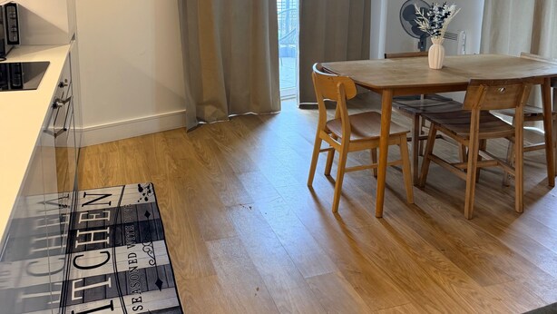 A modern living room with a wooden dining table, a sofa, a kitchen area, and a view of a building through large windows. - Cozy 2-bed Apt, Sleeps 4 Near Jewellery Quarter (Birmingham)