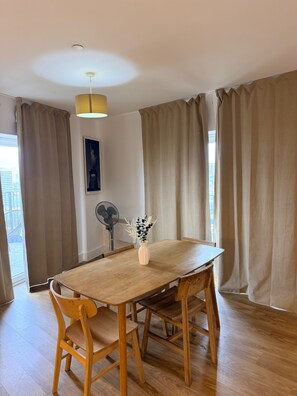 Un coin repas avec une table et des chaises en bois, un vase rempli de fleurs, un ventilateur de plafond et de grandes rideaux.