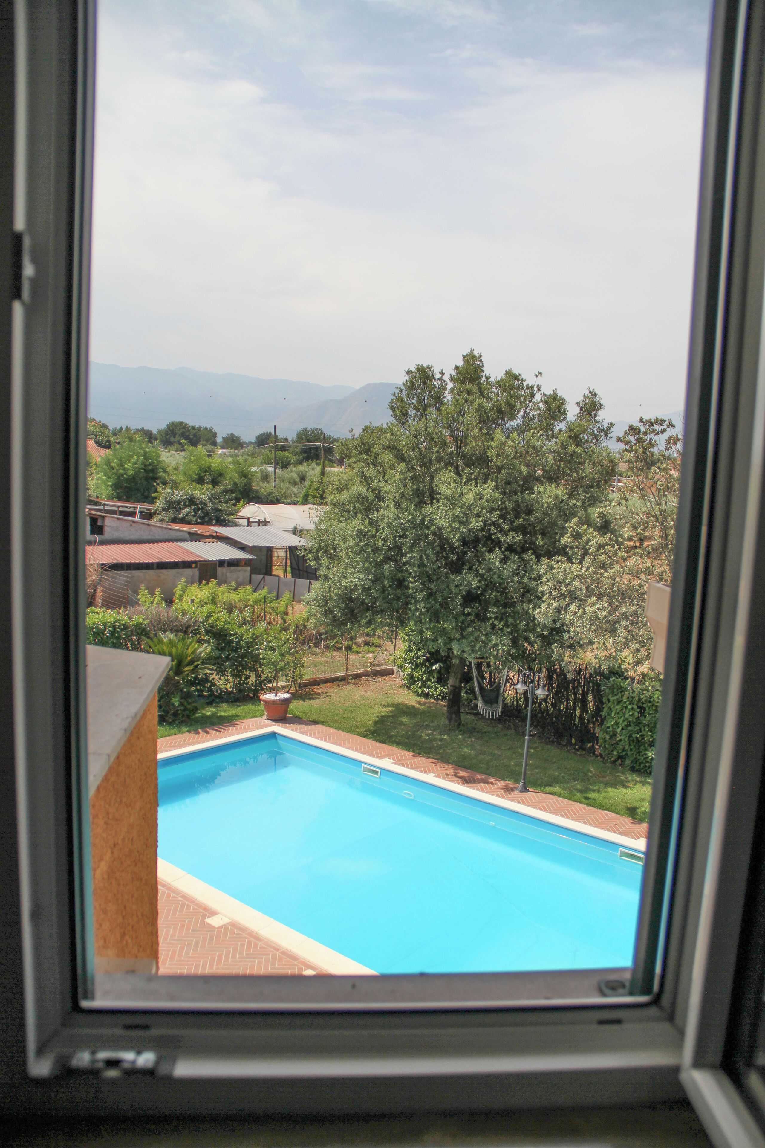 A view of a swimming pool through a window, surrounded by greenery and buildings.