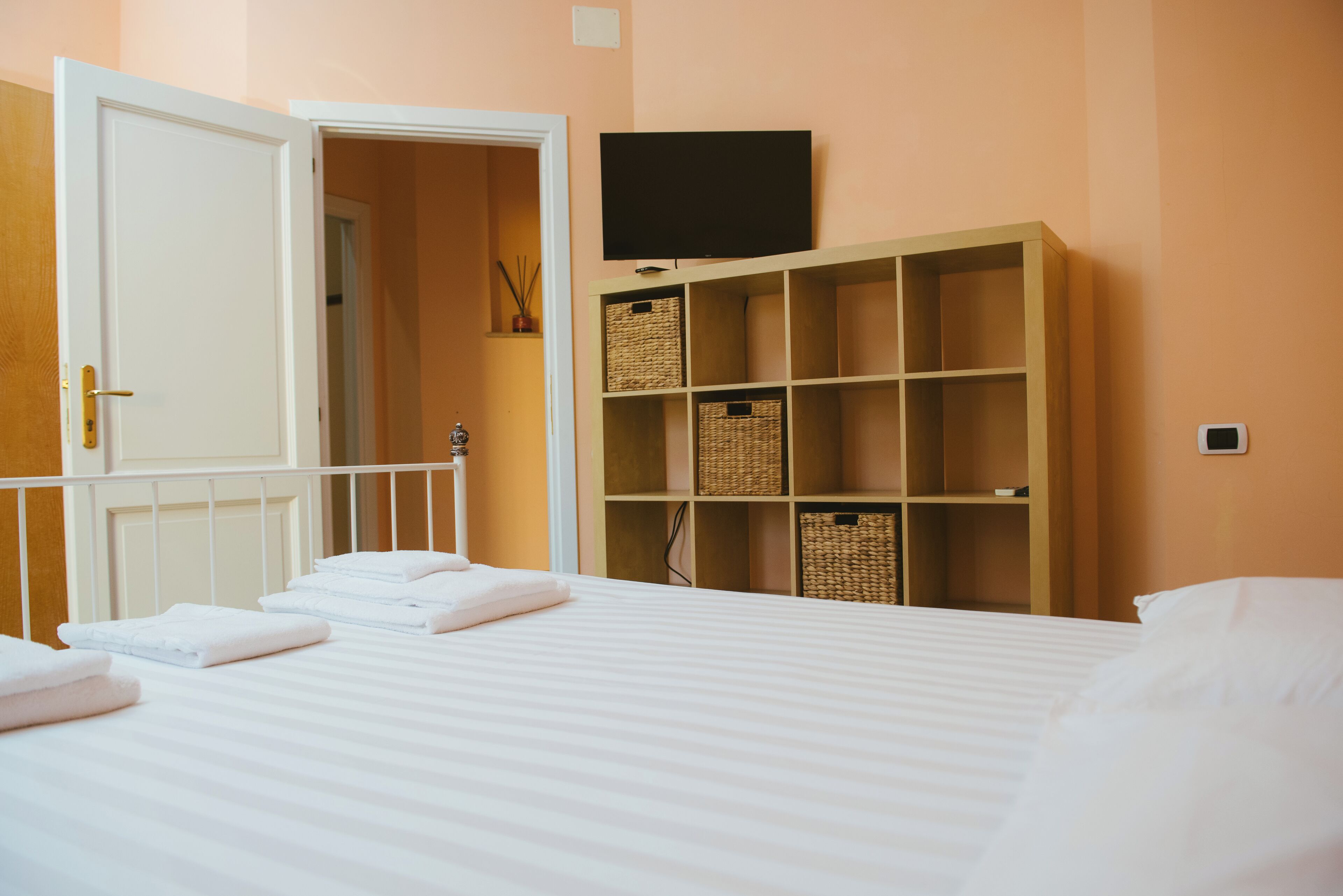 Una cama bien hecha con sábanas y toallas blancas, un televisor montado en la pared y una estantería de madera con cestas.