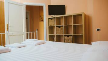Una cama bien hecha con sábanas y toallas blancas, un televisor montado en la pared y una estantería de madera con cestas.