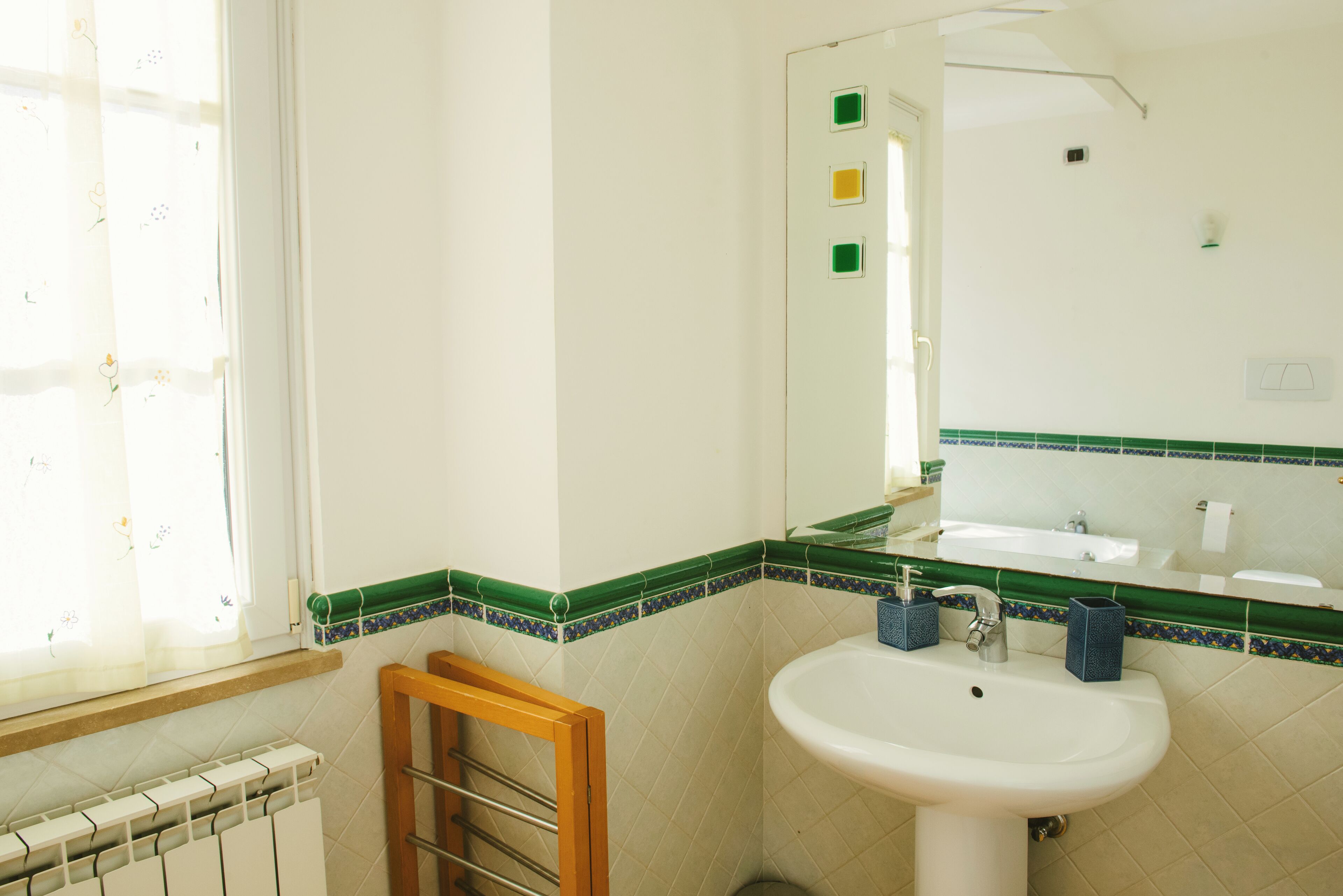 A bathroom with a white sink, green and blue tiled backsplash, a wooden towel rack, and a window with sheer curtains.