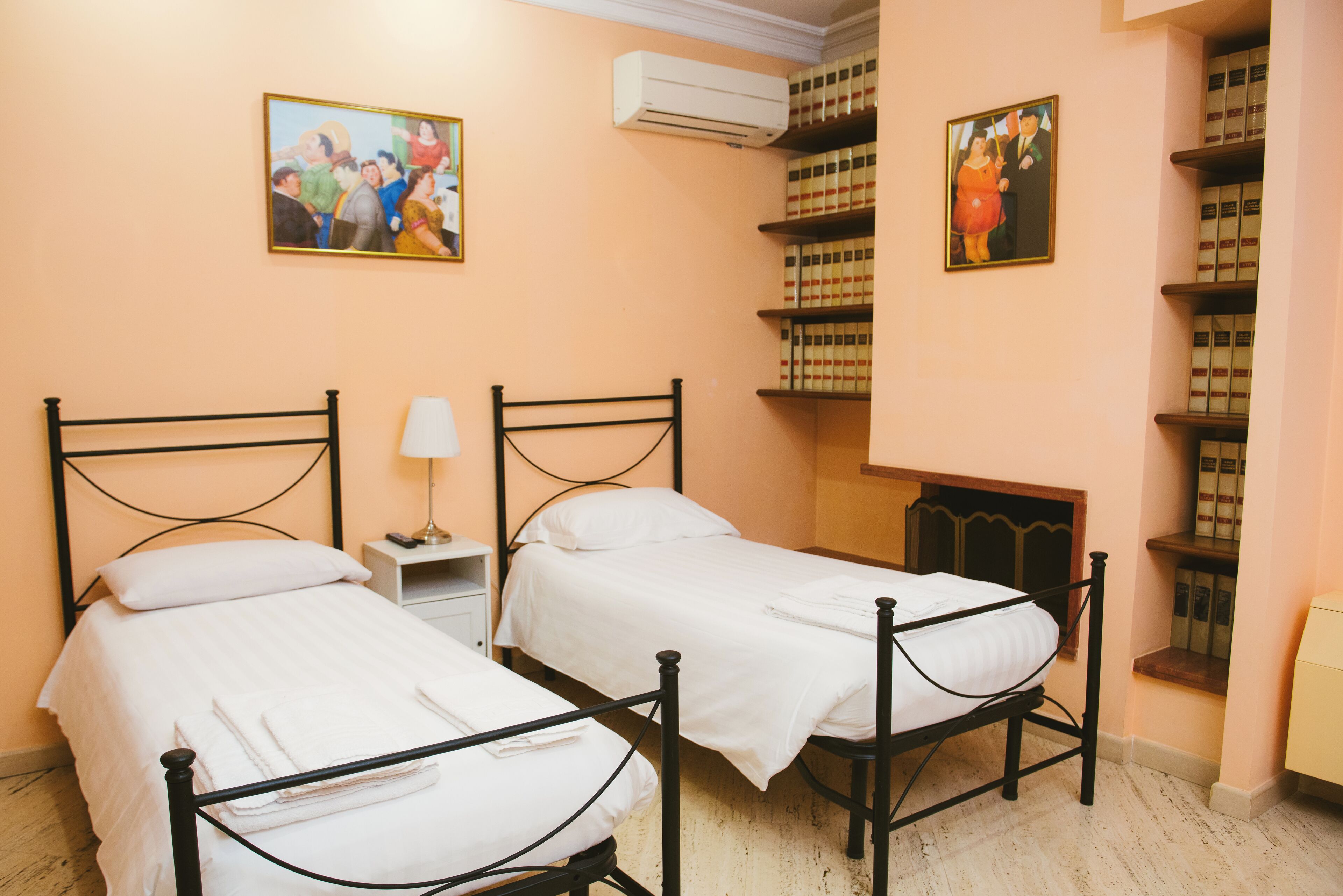 A room with two single beds, a bedside table with a lamp, a bookshelf, and a painting on the wall.